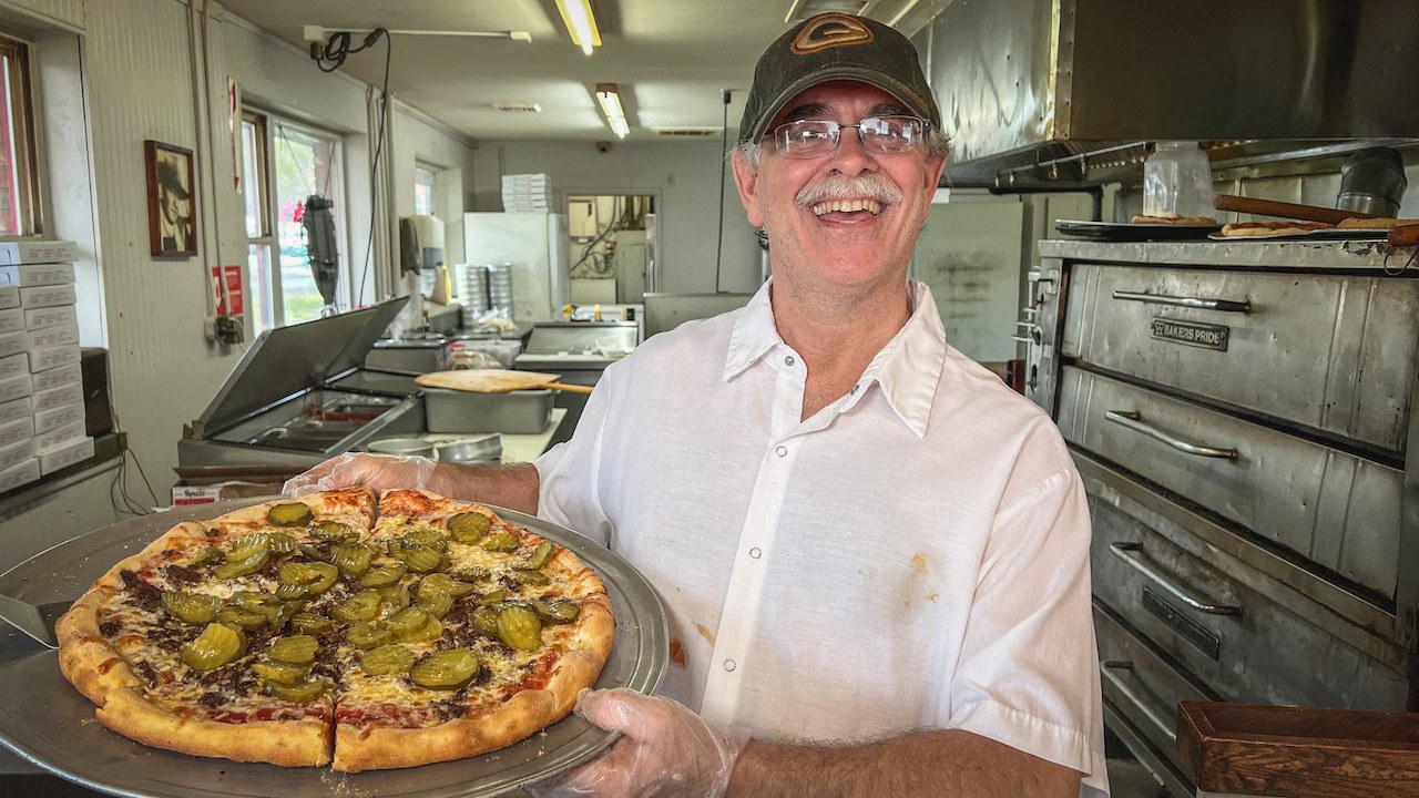 Robbie T holding a fresh cheeseburger pizza at his Jamesville shop